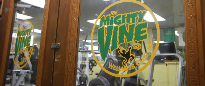 Mighty Vine Wellness Club logo displayed on a glass door with gym equipment visible inside.
